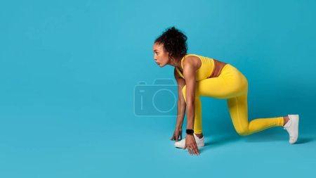 Téléchargez les photos : Vue latérale de la jeune femme noire en vêtements de sport jaune vif, posant en toute confiance en position de départ accroupie, en regardant l'espace de copie sur fond bleu studio. Bien-être et vie sportive. Panorama - en image libre de droit