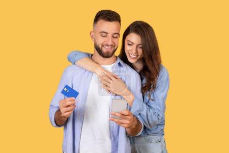 Foto de Pareja alegre compartiendo momento de comercio electrónico, hombre con tarjeta de crédito y mujer abrazándolo mientras ambos miran el teléfono inteligente, compras en línea, fondo amarillo - Imagen libre de derechos