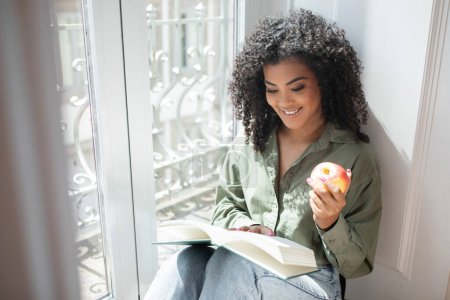 Téléchargez les photos : Joie de lire. Heureuse femme brésilienne millénaire se livre dans un livre best-seller, lit par la fenêtre tenant des pommes, profiter de son week-end détendu dans une maison confortable. Espace de copie - en image libre de droit