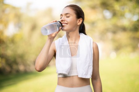 Foto de Una joven europea alegre sacia su sed bebiendo agua de una botella transparente, ojos cerrados de satisfacción, con una toalla blanca alrededor de su cuello en un parque soleado, al aire libre - Imagen libre de derechos