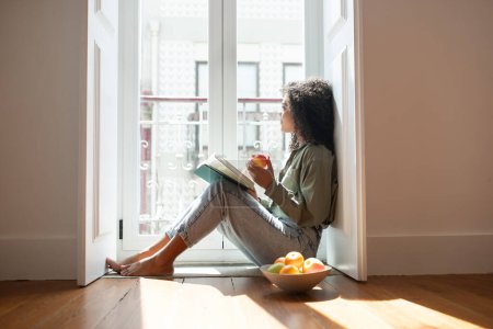 Téléchargez les photos : Lecteur moderne. Dame brésilienne absorbé dans un livre de lecture sur le sol dans un appartement confortable à l'intérieur, manger des fruits et apprendre la littérature par la fenêtre à la maison. Vue latérale - en image libre de droit