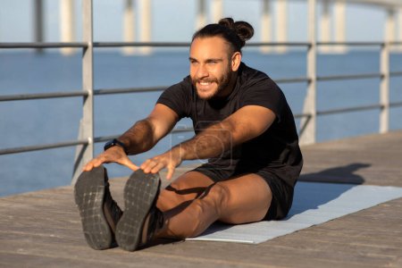 Photo for Happy athletic guy in sportswear demonstrates seated forward bend on mat, working out at seaside outdoor, showcasing flexibility and promoting exercise for wellbeing and fit lifestyle - Royalty Free Image