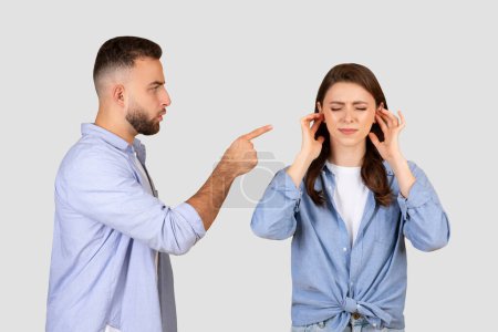 Téléchargez les photos : L'homme irrité pointe du doigt la dame en faute tandis que la femme en détresse couvre ses oreilles, toutes deux engagées dans un conflit sur fond de studio gris neutre - en image libre de droit
