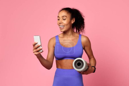 Fitness and tech. Athletic African American woman in purple activewear holds smartphone and foam roller, websurfing sporty apps, combining workout and online browsing on pink studio background