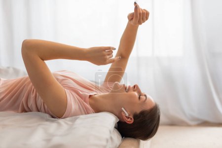 Photo for Relaxed young woman wearing earbuds while lying on her back in bed, smiling and raising hands, against soft light bedroom interior, side view, free space - Royalty Free Image