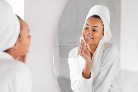 Foto de Señora negra después de la ducha se encarga de su piel utilizando productos cosméticos, aplicándolos con almohadilla de algodón mirando en el espejo en el moderno baño interior. Concepto de cuidado facial femenino - Imagen libre de derechos