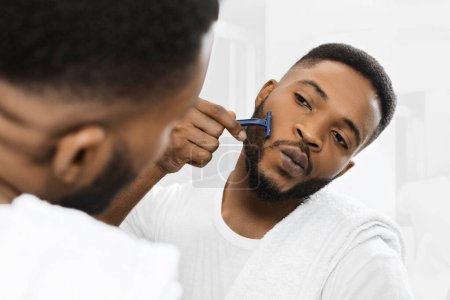 Téléchargez les photos : Afro homme rasant sa barbe en regardant dans le miroir à la salle de bain. Concept de soins de la barbe - en image libre de droit