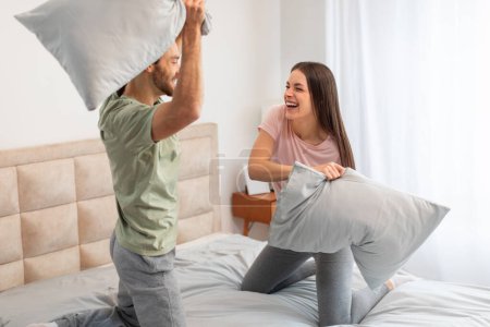 Riendo joven pareja que participan en juguetona pelea de almohadas en su cama, creando un ambiente animado y alegre en un entorno de dormitorio luminoso, disfrutando del tiempo juntos en la mañana
