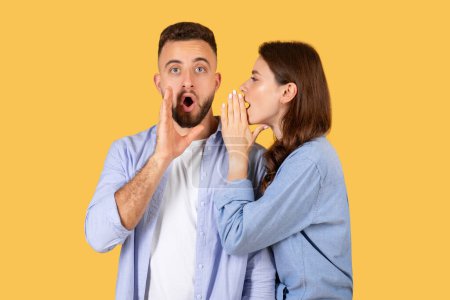 Surprised man listens with open mouth as woman whispers in his ear, both in casual attire against yellow background, sharing secret or news