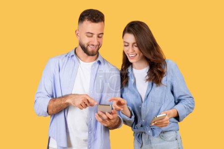 Foto de Hombre y mujer alegres en ropa casual intercambian contenido emocionante en sus teléfonos, disfrutando de un momento de interacción digital sobre un fondo amarillo cálido - Imagen libre de derechos