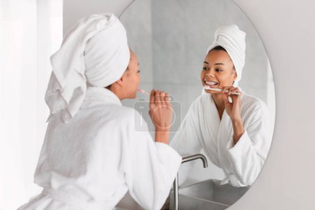 Téléchargez les photos : Afro-américaine dame pratique sa routine dentaire quotidienne brossant les dents à la salle de bain domestique à l'intérieur, la promotion de saines habitudes dans l'intérieur de la maison moderne, debout dans le peignoir près du miroir - en image libre de droit