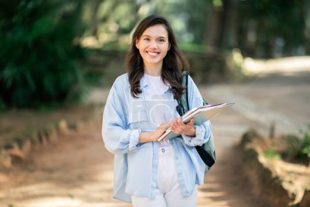 Téléchargez les photos : Une jeune étudiante joyeuse au sourire éclatant marche sur un chemin boisé du campus, tenant un manuel scolaire et portant une veste décontractée en denim sur une chemise blanche, avec un sac à dos pendu sur son épaule - en image libre de droit