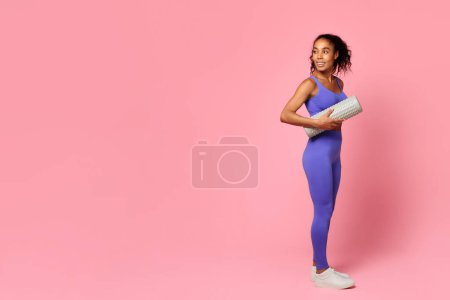 Téléchargez les photos : Mode de vie sain. Athlétique dame noire en violet sportswear tenant rouleau de mousse, représentant concept d'entraînement et de motivation, debout toute la longueur sur fond de studio rose. Espace libre - en image libre de droit