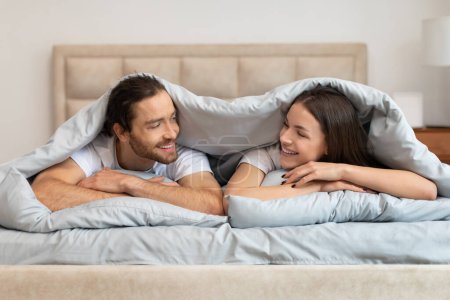 Foto de Alegre pareja comparte momento de luz bajo la manta, charlando con amplias sonrisas, mostrando sentido de intimidad y comodidad en su entorno dormitorio - Imagen libre de derechos