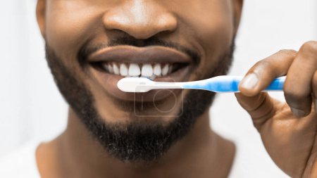 Téléchargez les photos : Jeune homme afro souriant avec brosse à dents nettoyant les dents dans la salle de bain. Concept sain de blanchiment des dents - en image libre de droit
