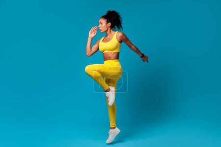 Fitness banner. African American sportswoman in yellow activewear demonstrates strength and cardio exercise, doing elbow to knee standing pose with determination, over blue backdrop