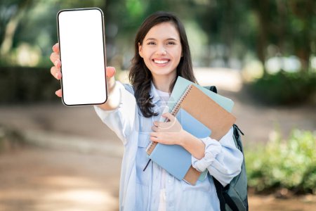 Photo for A cheerful caucasian young student lady shows her smartphone screen while holding textbooks, embodying a blend of technology and education in a vibrant campus setting, outdoor - Royalty Free Image