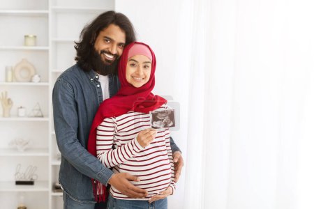 Foto de Embarazada pareja musulmana sosteniendo el ultrasonido del bebé mientras están parados en el interior del hogar juntos, amando a los cónyuges islámicos mostrando la imagen de sonografía de su hijo, disfrutando de la próxima paternidad, espacio para copiar - Imagen libre de derechos