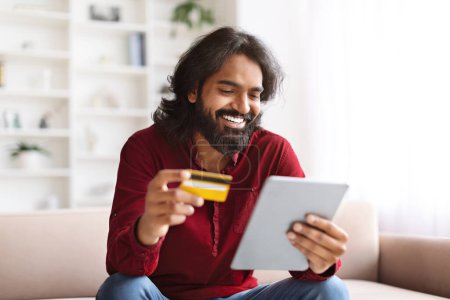 Foto de Alegre sonriente guapo joven indio sentado en el sofá en la sala de estar, utilizando tableta digital y tarjeta de crédito bancaria, ordenar comida en línea, compras en Internet desde casa, espacio para copiar - Imagen libre de derechos
