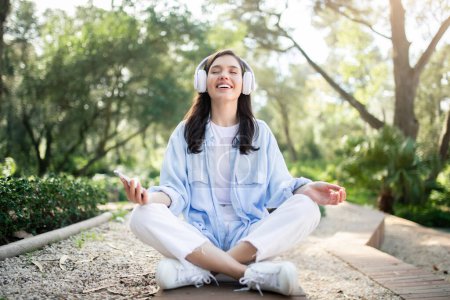 Téléchargez les photos : Une femme millénaire caucasienne heureuse et sereine profite de la méditation dans un parc ensoleillé, écouteurs et smartphone à la main, respirant la paix et la détente moderne, profiter du temps libre, en plein air - en image libre de droit