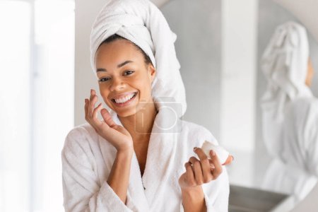 Téléchargez les photos : Portrait de dame noire joyeuse posant avec pot de crème comme elle hydrate son visage dans l'intérieur de la salle de bain, debout dans un peignoir blanc et une serviette enveloppée autour de la tête, l'application de produits de beauté - en image libre de droit