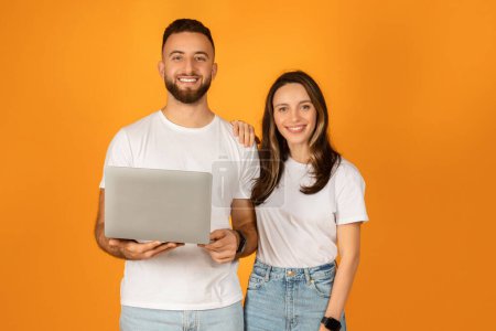 Photo for Cheerful young couple with a man holding a laptop and woman standing close, both dressed in white t-shirts and jeans, exuding confidence and happiness against a plain orange background - Royalty Free Image