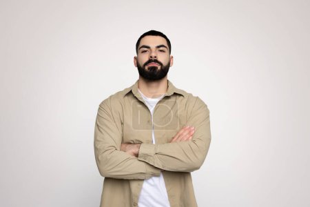Téléchargez les photos : Homme arabe millénaire rassuré avec une barbe soignée bras debout croisés dans une veste beige et t-shirt blanc, exsudant confiance et professionnalisme dans un contexte gris - en image libre de droit