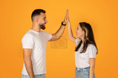 Photo for Happy young couple giving a high-five, celebrating success with joyous expressions, both dressed in white t-shirts and blue jeans against an orange background, symbolizing teamwork and achievement - Royalty Free Image