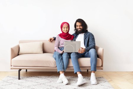 Photo for Portrait Of Romantic Muslim Spouses Relaxing With Laptop At Home, Happy Young Arabic Couple Embracing And Using Computer, Resting On Couch In Living Room, Browsing Internet Or Shopping Online - Royalty Free Image