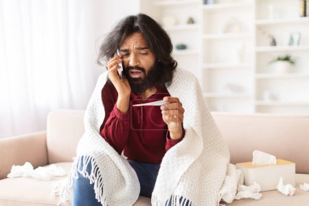 Photo for Unhealthy millennial indian guy covered in warm blanket sitting on couch at home, hold electronic thermometer in his hand, checking body temperature. Sick eastern man suffering from fever, copy space - Royalty Free Image