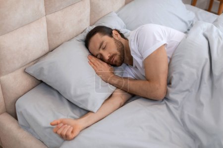 Téléchargez les photos : Homme en t-shirt blanc est sonore endormi, mettant en valeur la tranquillité de se reposer dans un lit confortable avec des oreillers moelleux et des draps soyeux, évoquant la sérénité, vue ci-dessus - en image libre de droit