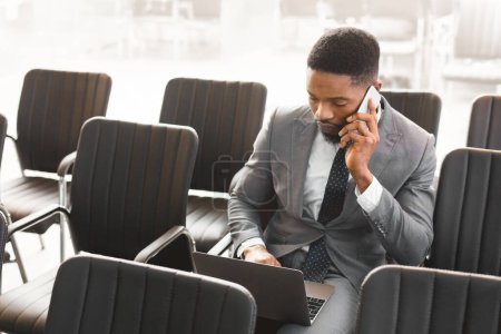Téléchargez les photos : Homme d'affaires afro concentré travaillant sur ordinateur portable dans la salle de conférence vide, ayant parlé au téléphone - en image libre de droit