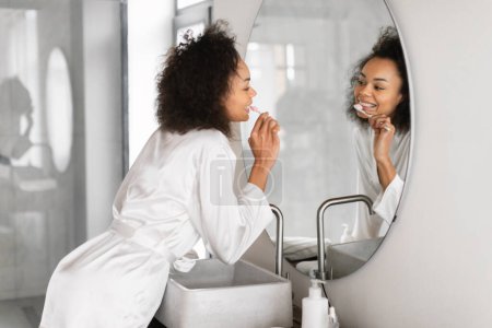 Téléchargez les photos : Jeune femme noire se brosse les dents devant le miroir de salle de bain à l'intérieur, embrasse la routine dentaire du matin en prenant soin de la propreté des dents et un sourire parfait, se préparer le matin - en image libre de droit