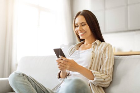 Photo for Cheerful young woman browsing her smartphone while comfortably lounging on couch at home, happy millennial female using mobile phone while relaxing on sofa in living room interior, copy space - Royalty Free Image