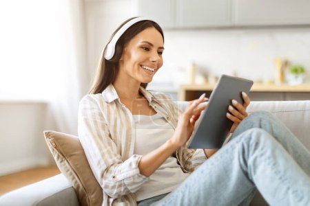 Photo for Cheerful young woman relaxing on couch, wearing headphones and holding digital tablet, smiling millennial female enjoying music, watching content or making video call, resting in living room at home - Royalty Free Image