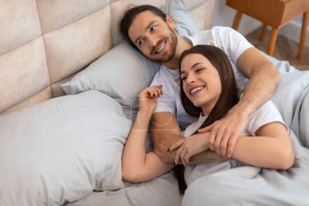 Photo for Cheerful couple in white t-shirts enjoys light-hearted moment, lying in bed with bright smiles, exuding warmth and intimacy in their bedroom, above view, free space - Royalty Free Image