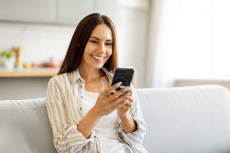Foto de Sonriendo hermosa mujer escribiendo mensaje en su teléfono inteligente mientras está sentada en el sofá en casa, feliz joven europea femenina usando el teléfono móvil para la comunicación o compras en línea, Copiar el espacio - Imagen libre de derechos