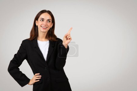 Foto de Empresaria milenaria caucásica optimista con una sonrisa radiante, vistiendo una chaqueta negra, apuntando casualmente hacia arriba, sugiriendo una tendencia o idea positiva, con un fondo claro - Imagen libre de derechos