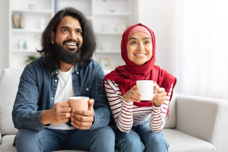 Téléchargez les photos : Couple musulman heureux boire du café et se détendre sur le canapé dans le salon, conjoints arabes joyeux regarder la télévision à la maison et profiter de boissons chaudes, passer le week-end ensemble, gros plan - en image libre de droit