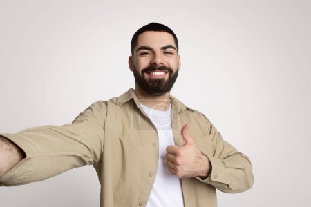 Téléchargez les photos : Joyeux homme arabe barbu millénaire prenant un selfie d'une main tout en donnant un pouce vers le haut avec l'autre, portant une chemise beige décontractée sur un t-shirt blanc, respirant la confiance et la convivialité - en image libre de droit