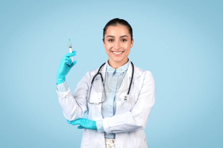 Smiling female doctor confidently holding syringe, wearing sterile gloves and white lab coat, ready for patient care on light blue background