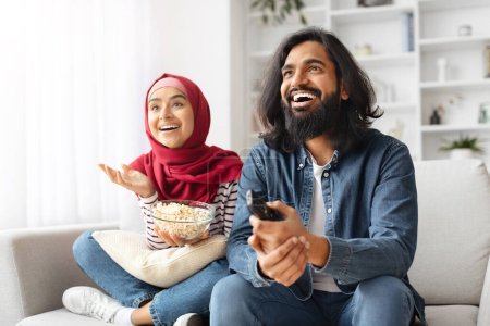 Foto de Alegre pareja musulmana disfrutando de la película divertida en la televisión mientras se relaja en el sofá en casa, cónyuges islámicos felices riendo juntos y comiendo palomitas de maíz, descansando en la sala de estar acogedora brillante, espacio libre - Imagen libre de derechos