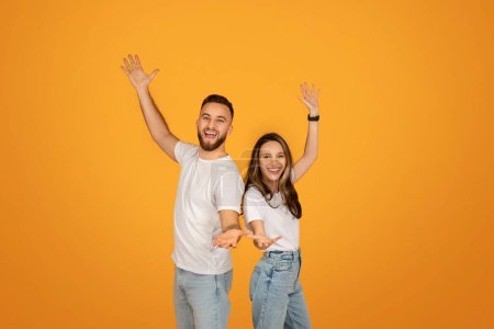 Téléchargez les photos : Joyeux homme et femme caucasiens en hauts blancs et en jeans bleus lèvent les bras haut dans un geste ludique et accueillant, rayonnant de bonheur sur un fond orange chaud - en image libre de droit
