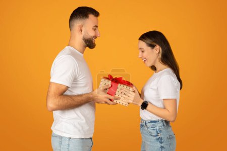 Photo for A cheerful caucasian millennial man in a white t-shirt gives a wrapped gift with a red bow to a delighted woman in casual attire against a vibrant orange background, studio - Royalty Free Image