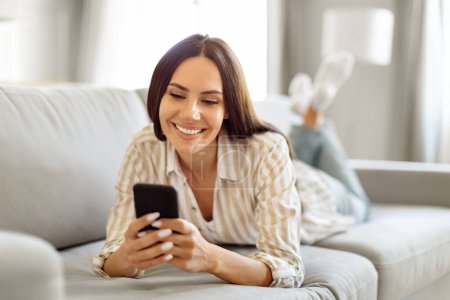 Photo for Happy young european lady messaging on smartphone while relaxing at home, smiling brunette woman lying on couch in living room interior, enjoying online chat on her phone, copy space - Royalty Free Image