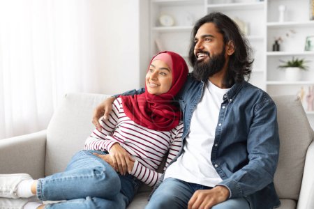 Photo for Happy young muslim couple in casual wear sitting closely on couch and smiling together, romantic arabic man and woman in hijab enjoying peaceful moment at home, resting on sofa in their living room - Royalty Free Image