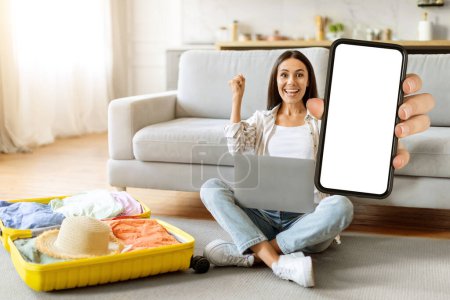 Photo for Hot traveling offer. Happy young woman using laptop and showing blank smartphone with blank screen, excited female sitting on floor at home, packing suitcase, demonstrating mockup for advertisement - Royalty Free Image