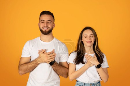 Téléchargez les photos : Reconnaissant jeune couple caucasien avec les yeux fermés et les mains sur les cœurs, exprimant des émotions sincères et de l'appréciation, portant des t-shirts et des jeans blancs, sur un fond orange chaud - en image libre de droit