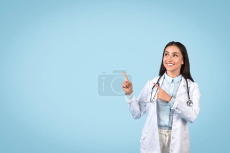 Photo for Joyful female doctor pointing upwards at free space with both hands, showing excitement, in white lab coat with stethoscope, against serene blue background, medical ad - Royalty Free Image