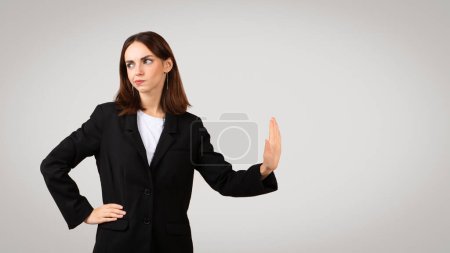 Photo for Distrustful caucasian millennial businesswoman in a black suit jacket showing a stop hand sign, with a skeptical expression, indicating caution or refusal, against a plain background - Royalty Free Image